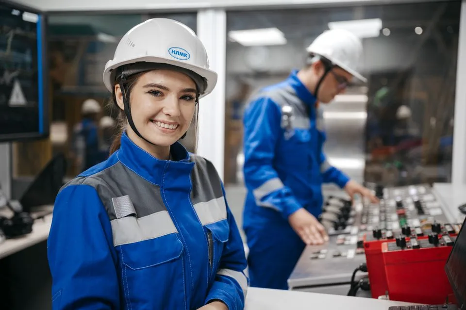 Lächelnde Fachkraft im Vordergrund mit Arbeitskleidung und Helm mit geschlossenem Kinnriemen, vor Kollegen in einer industriellen Leitwarte