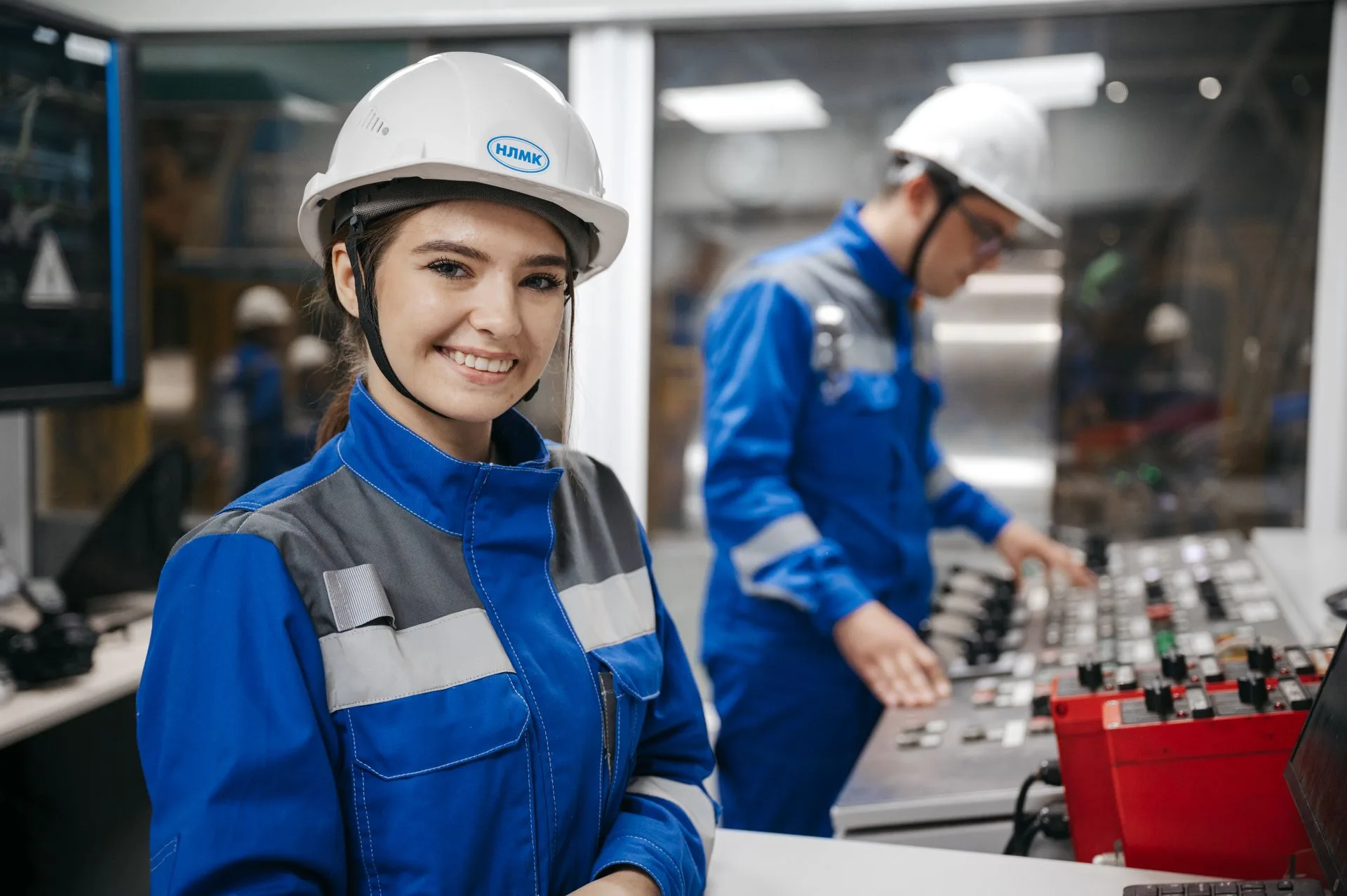 A smiling professional in the foreground, wearing a uniform and a hard hat with the chin strap fastened, with colleagues behind in an industrial control room