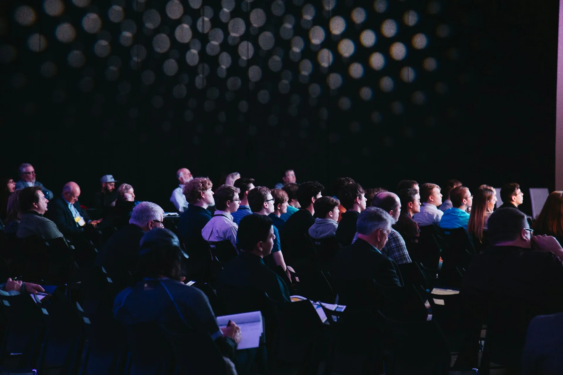 An audience seated in an auditorium watching a presentation; some people are taking notes.