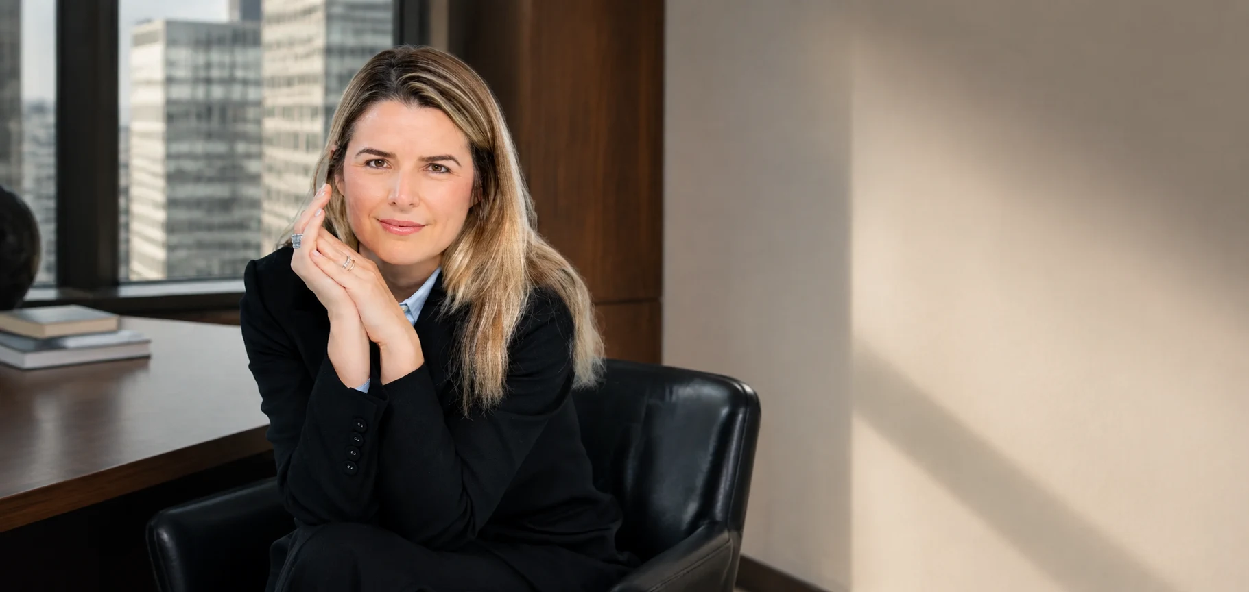 Andreza Araújo sentada, de blazer preto e camisa azul, em composição de escritório em Nova York